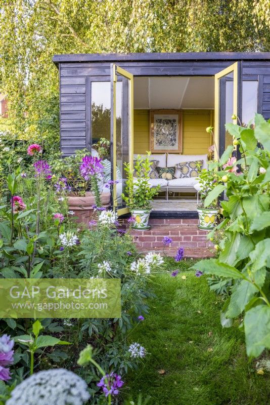 Black timber summer house with open doors showing interior and pots of lemon trees on steps outside and borders of annuals and Dahlias