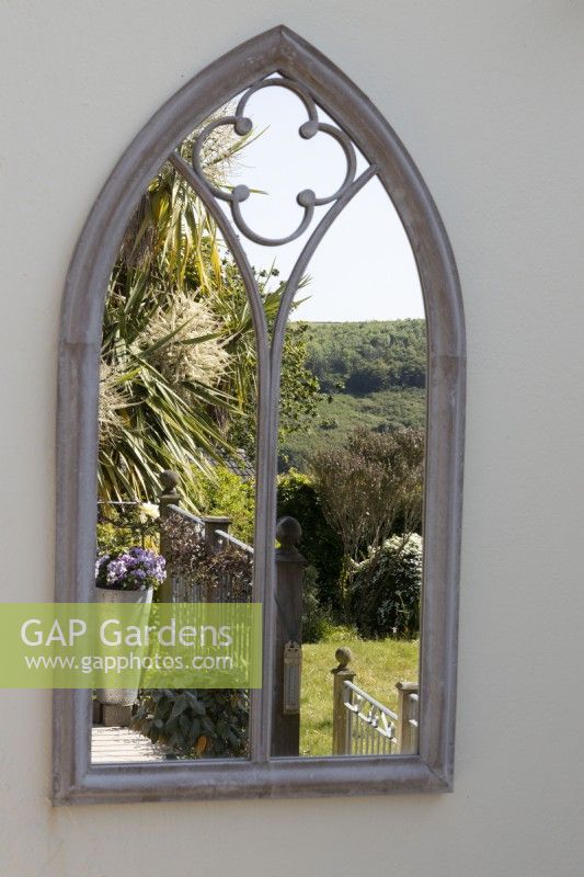 An arched gothic style mirror on a cream wall reflects a balcony, with Cordyline australis and far reaching countryside view. 