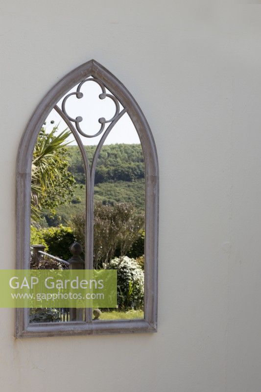 An arched gothic style mirror on a cream wall reflects a balcony, with Cordyline australis and far reaching countryside view. May. 