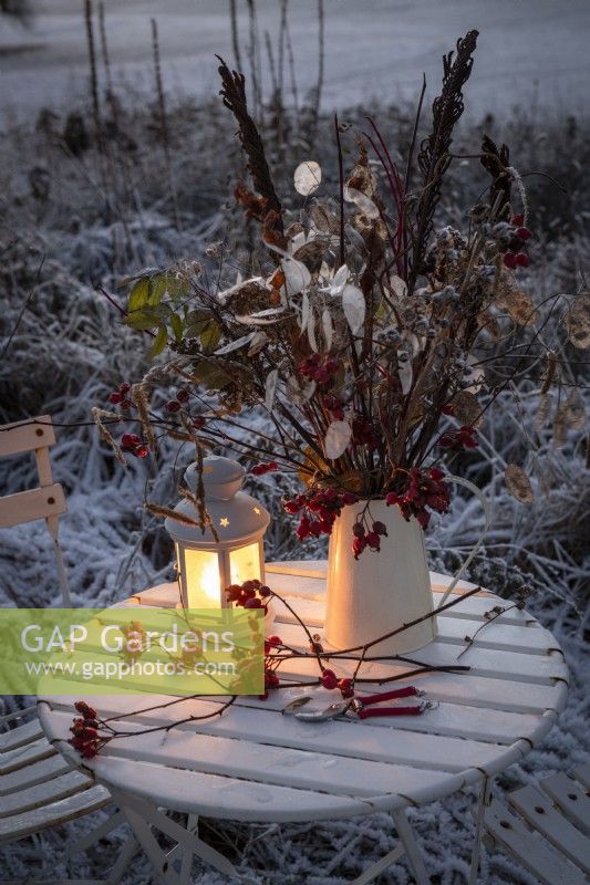 Winter display of frosted honesty seedheads, dogwood stems, ostrich fern fronds and rose hips in a metal jug on a bistro table. Metal lantern with lit candle, Felco secateurs and metal chairs. Snow covered garden in background.