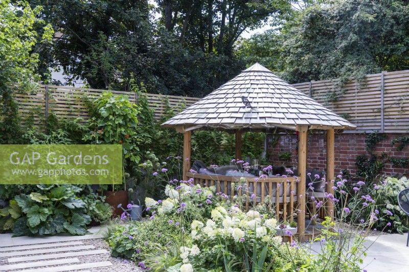 Round wooden gazebo with tile roof surrounded by mixed planting in garden borders