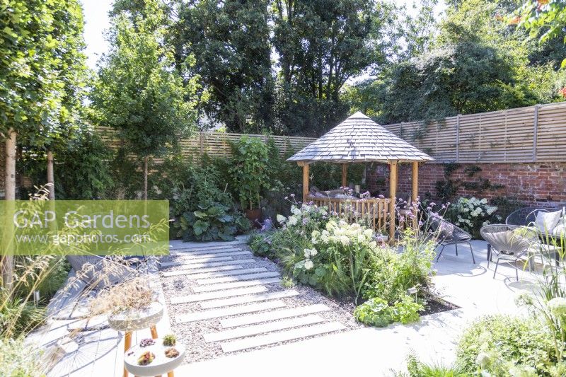 Garden with stone tile patio and staggered path with mixed planting beds and borders, seating areas and a round wooden gazebo with a tile roof