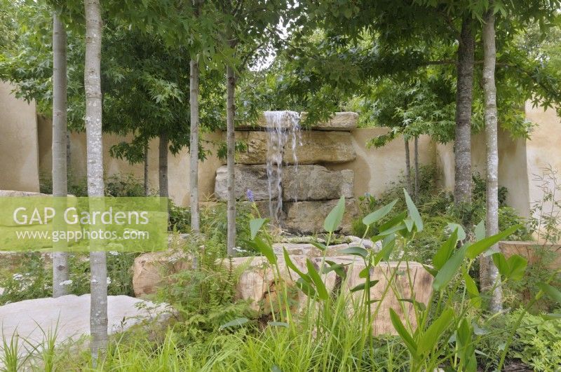 Woodland plants and trees frame a waterfall cascading over slabs of rock providing a dynamic focal point.  America's Wild, presented by Trailfinders  and  Visit The USA, RHS Hampton Court Palace Garden Festival 2023.  