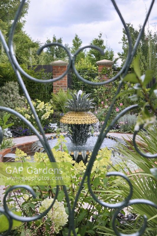 View through a green wrought iron gate of a formal garden based on the historic courtyard gardens of Charleston, South Carolina, with a pineapple water feature representing the famous Charleston waterfront fountain.  Explore Charleston - Welcome to Charleston, RHS Hampton Court Palace Garden Festival 2023.  