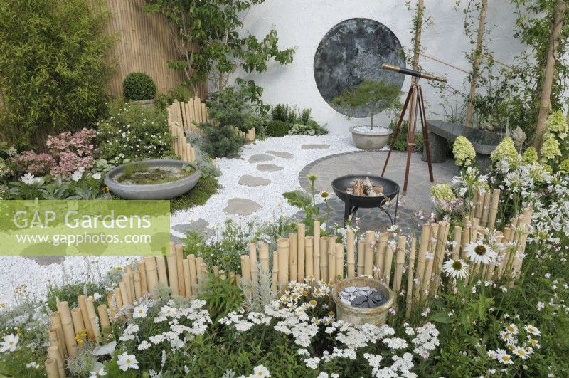 A courtyard garden with a repetition of circular shapes and a winding gravel path leading to a round mottled antique mirror on the back wall - reminiscent of an Oriental moon gate.  Plants including Echinacea purpurea 'White Swan', Achillea millefolium 'White Beauty', Cosmos bipinnatus 'Purity' with contrasting silver green foliage along undulating bamboo fences. The Lunar Garden, RHS Hampton Court Palace Garden Festival 2023.  