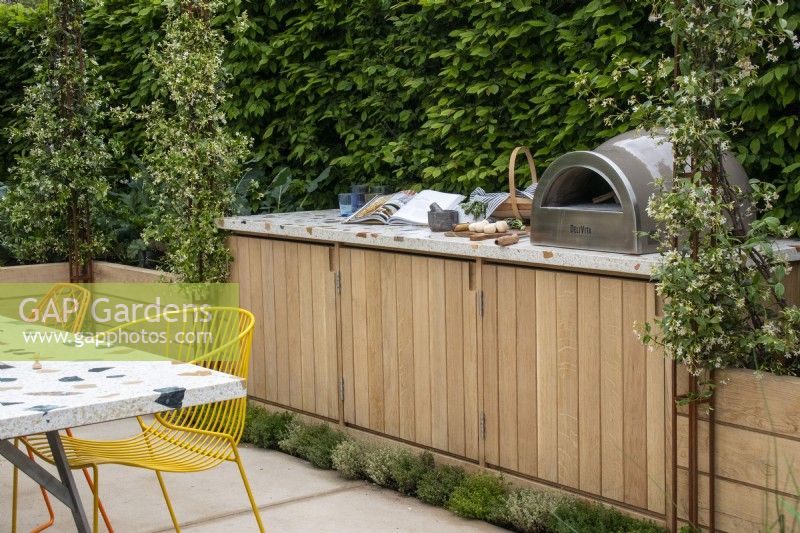 An outdoor kitchen backed by a hedge and will metal obelisks supporting flowering Trachelospermum jasminoides - The London Square Community Garden - Sanctuary Gardens - Designer James Smith - RHS Chelsea Flower Show 2023