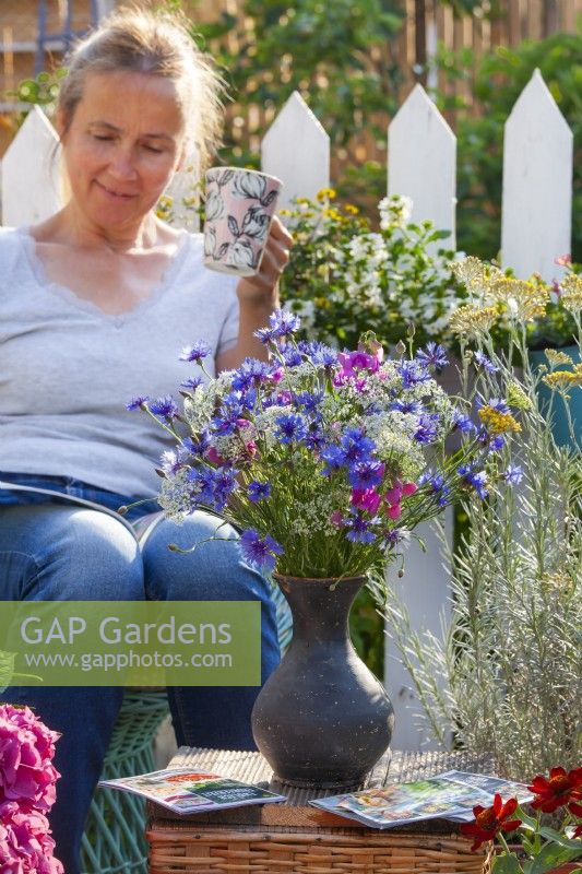 Flower bouquet with cornflowers, wild carrots and sweet peas, woman in the background enjoys reading magazine and drinking tea.