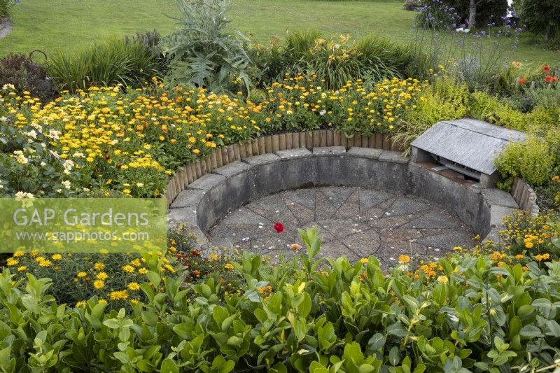 Journey to the Sun, the sunken barbecue area, with circular seating around the edge, amongst a profusion of yellow and orange planting. Harbour Lights, Devon NGS garden. July. 