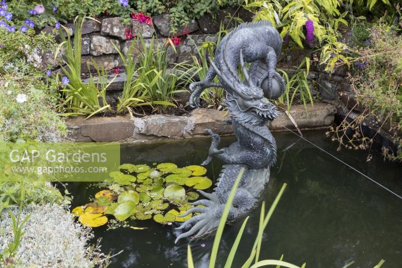 A large copper dragon sculpture, Chinese style, in a pond setting with a stream of water spraying from the mouth. Summer