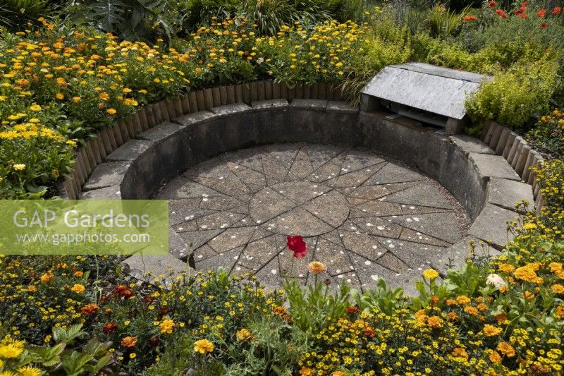 Journey to the Sun, the circular, sunken barbecue area amongst a profusion of yellow and orange planting, with a paved sun shape in the middle and seating around the edges. Harbour Lights, Devon NGS garden. July. 