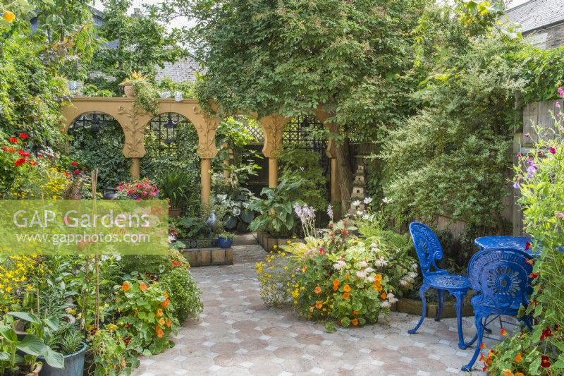 Small, paved courtyard garden styled with a Moroccan theme. Colourful bedding plants in containers with hostas, nasturtiums, cosmos, bidens and begonias. Blue painted metal patio table and chairs. 
July.