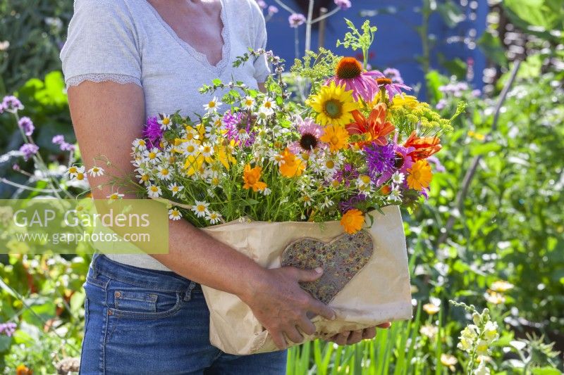 Woman holding paper bag with harvested edible flowers including sunflowers, monarda, coneflowers, hemerocallis, pot marigolds and chamomile.