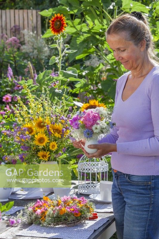 Woman arranging outdoor table with wreath and bouquets of flowers in vases. Flowers in bouquets are Hydrangea, sweet peas, Ammi majus, Helianthus and Nigella while wreath is made of Clematis seed heads, Dahlia and Rudbeckia.