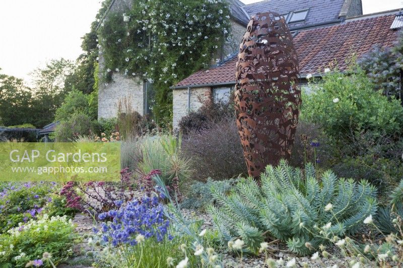 Gravel garden by house with Hylotelephium telephium (Atropurpureum Group) 'Purple Emperor' and Verbena officinalis var. grandiflora 'Bampton' complementing the sculpture and contrasting with Euphorbia rigida.