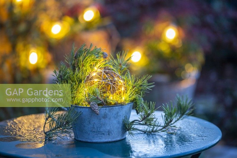 Wicker ball wrapped in lights in a metal pot with Pine sprigs on a metal table with a thin layer of ice