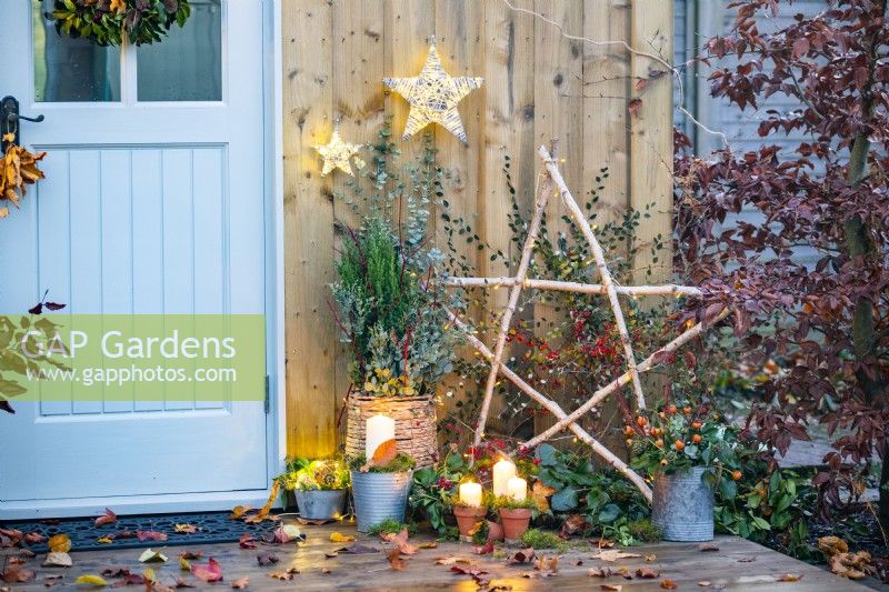 Candles in terracotta pots and one in a metal bucket with moss in front of Birch star, wicker pot containing Juniper, Cornus sticks and Eucalyptus sprigs with a trug of Ivy and wicker light up stars hanging on the wall above