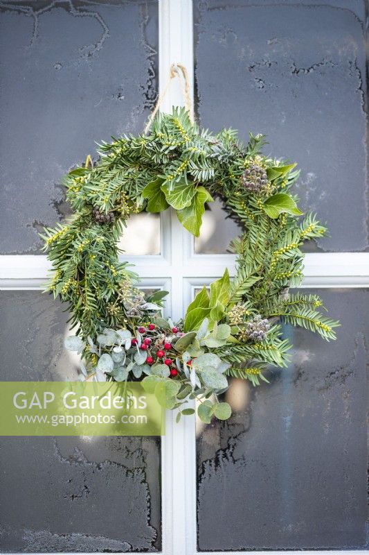 Wreath made of Yew, Ivy Hedera, Eucalyptus and Hawthorn berries hanging from door against frosted glass