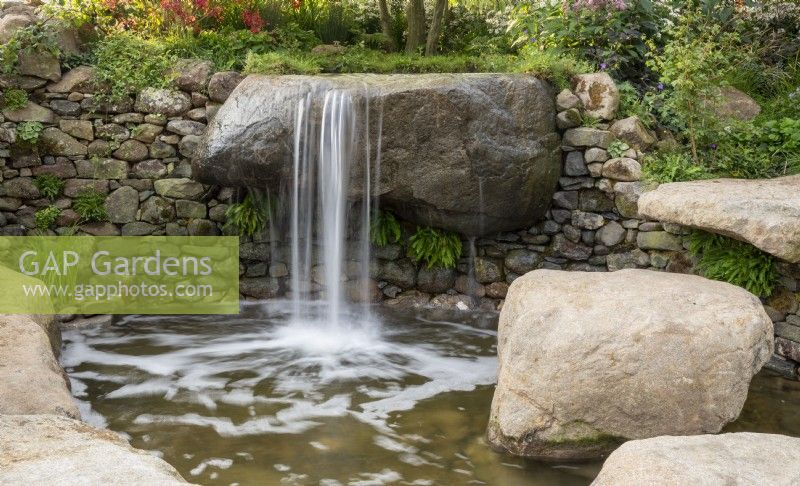 A waterfall over large stone boulders into a pond with Asplenium scolopendrium and damp loving plants growing out of a dry stone wall 
