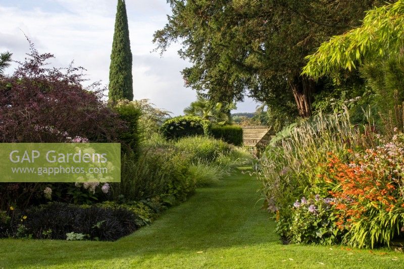 View down lawn past perennial borders towards the shade house at Bourton House Garden, Gloucestershire.