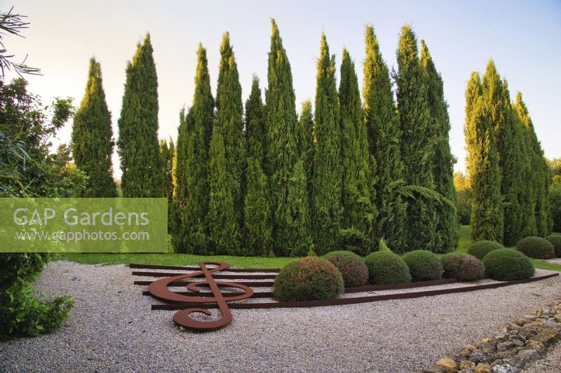 Art installation of The musical stave with treble clef sculpture and topiary bushes in the form of notes on the gravel in Mediterranean garden with rows of Cupressus Sempervirens or Italian Cypress trees on background. 
Italy, Tuscan Maremma, Orbetello
Autumn season, October
