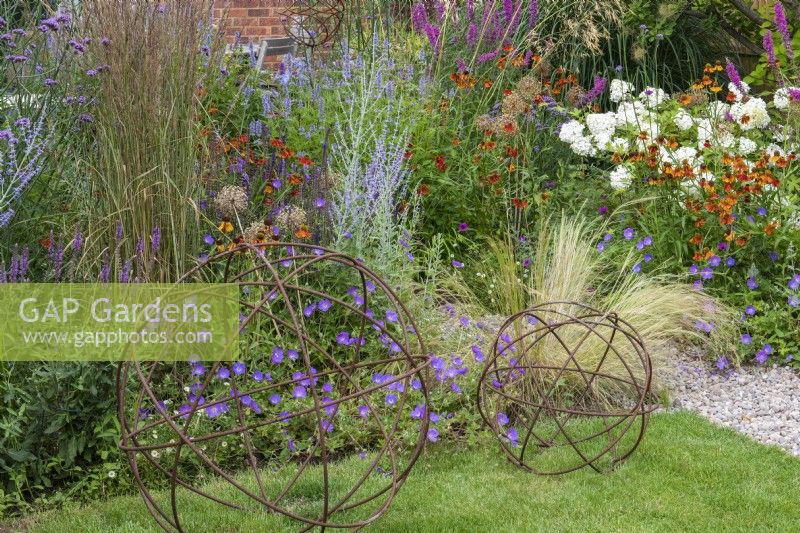 On the lawn, two rusty sphere decorations set against a backdrop of Geranium 'Azure Rush', Helenium 'Moerheim Beauty', Lythrum salicaria 'Fire Candle', Verbena bonariensis, hydrangea and feather reed grass.