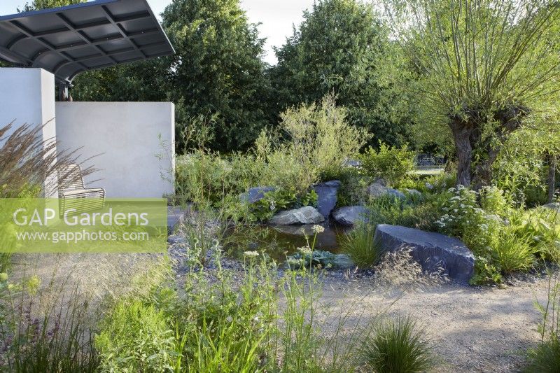 Cancer Research UK Legacy Garden. Seating area in the Pledge Pavillion. Pathway around rocky island border with Salix, hostas, grasses and Tanacetum. Summer.