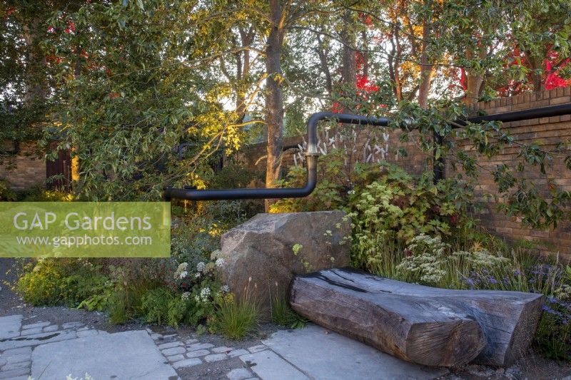 Urban garden lit up at night with mixed perennial planting - carved tree trunk bench seating area and a brick wall and reclaimed repurposed pipe sculpture runs through the garden