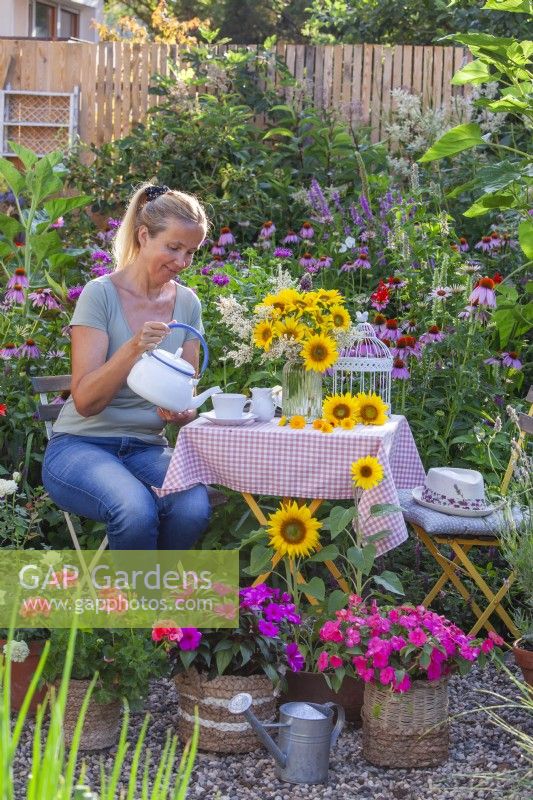 A woman enjoys tea time on a small terrace with pots of bedding flowers including Impatiens and Pelargonium.