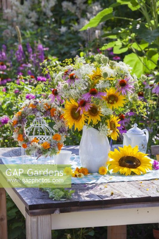 Wreath made of summer flowers hanging from bird cage and bouquet in enamel jug containing sunflowers, coneflowers, wild carrots and fennel.