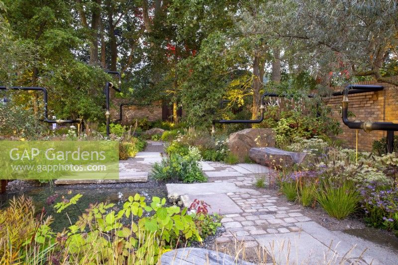 Reclaimed stone paving and mixed perennial in a new urban garden with benches, pond and pipe sculpture