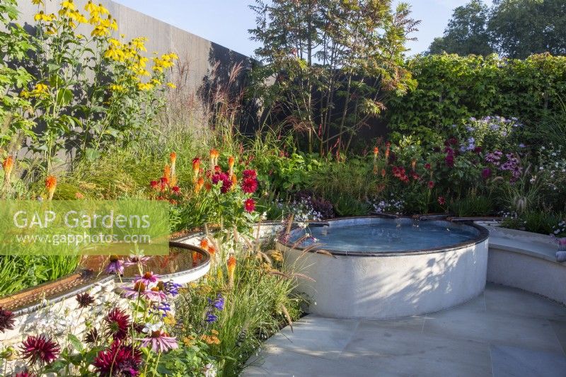 A courtyard garden with white paving slabs copper metal rill with water flowing into a raised small pool - mixed perennial planting of Echinacea purpurea, Dahlias, Kniphofia 'Papaya Popsicle' and ornamental grasses 
