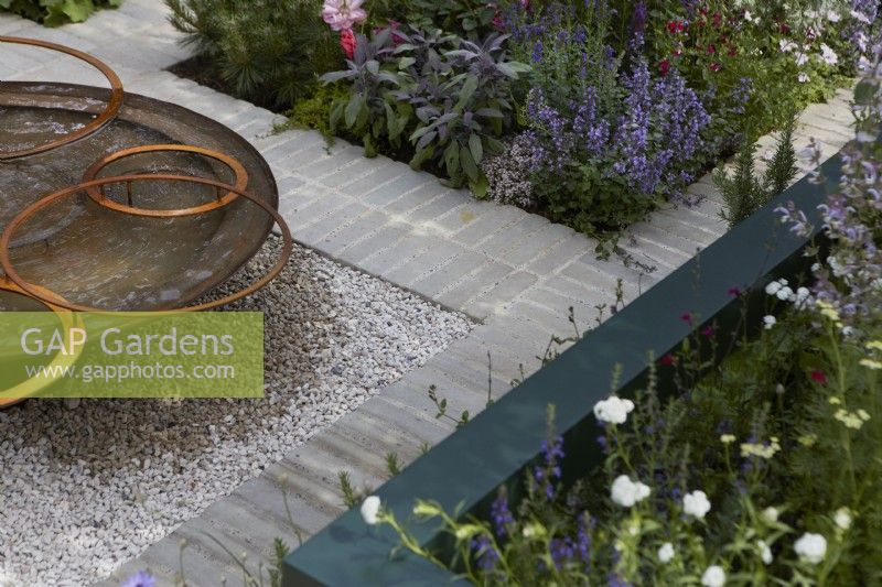 Landform Mental Wealth Garden. Designer: Nicola Hale. Detail of border edges with reclaimed circular metal water feature with rusty hoops. Summer.