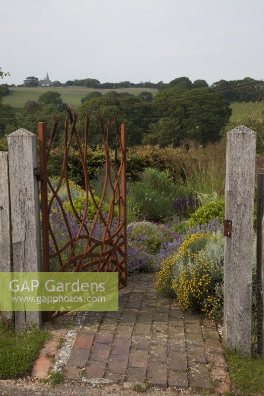 Drought tolerant garden filled with mediterranean plants. Bespoke Steel Gate allowed to rust, depicting corn and the fields, framing the view of the fields to Pett Church and entrance to The Jewel Garden -  Curved brick pathway. Nepeta 'Six Hills Giant' - Catmint,   Santolina chamaecyparissus ' Yellow Buttons', Euphorbia seguieriana subsp. niciciana , grasses including Calamagrostis brachytricha and Stipa gigantea, Geraniums, Buxus sempervirens balls - Box.
Gate Made by Jake Bowers The Thirsty Bear Forge Hastings