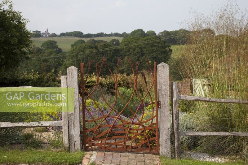 Drought tolerant garden filled with mediterranean plants. Bespoke Steel Gate allowed to rust, depicting corn and the fields, framing the view of the fields to Pett Church and entrance to The Jewel Garden. Plants include Euphorbia seguieriana subsp. niciciana and Stipa gigantea. Gate Made by Jake Bowers The Thirsty Bear Forge Hastings
