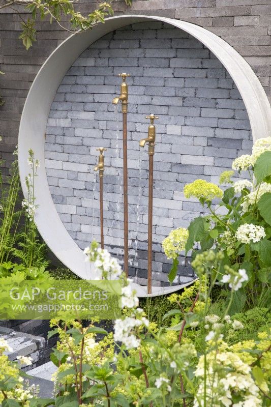 A circular water feature using stacked grey clay brick pavers and concrete surround against a wall with salvaged brass taps as water spouts and Hydrangea arborescens 'Annabelle'