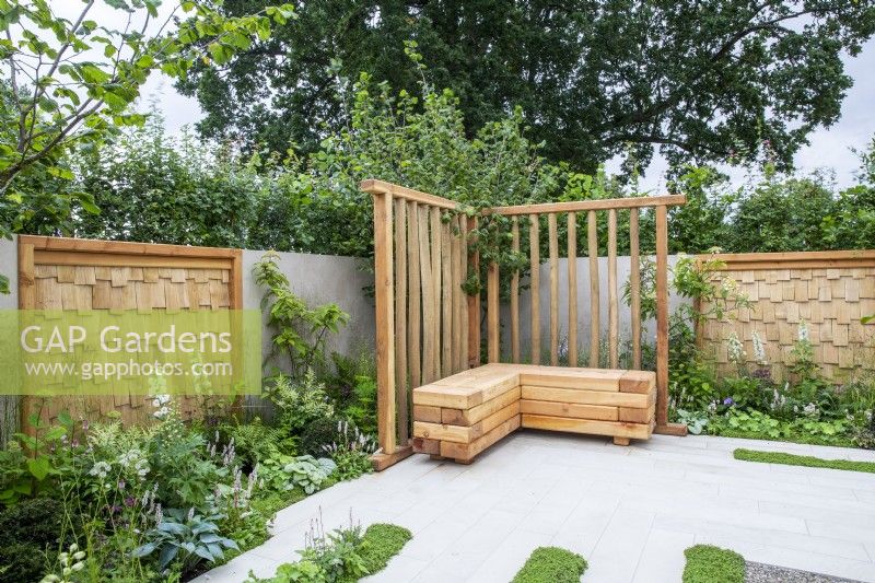 Modern contemporary patio area with grey stone paving, wooden bench seat and sweet chestnut screens on the walls, mixed perennial planting in white and green tones 