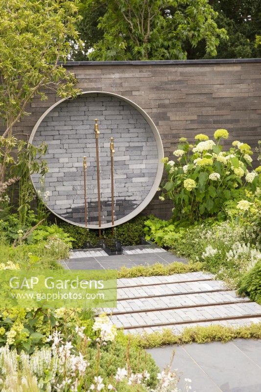 Garden wall made from stacked concrete slabs, circular water feature with brass taps, mixed perennial planting in white and green colours, Hydrangea arborescens 'Annabelle', grey clay brick pavers and stone paving