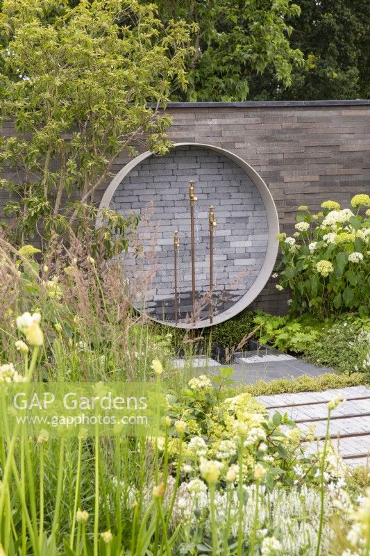 Garden wall made from stacked concrete slabs with a circular water feature with salvaged brass taps, mixed perennial planting in white and green colours