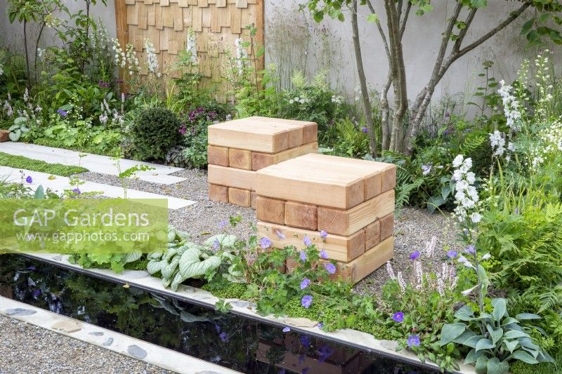 Wooden cube seats in a gravel garden surrounded by mixed perennial planting including Delphinium 'Guardian White', Brunnera macrophylla 'Jack Frost', Geranium wallichianum 'Azure Rush'