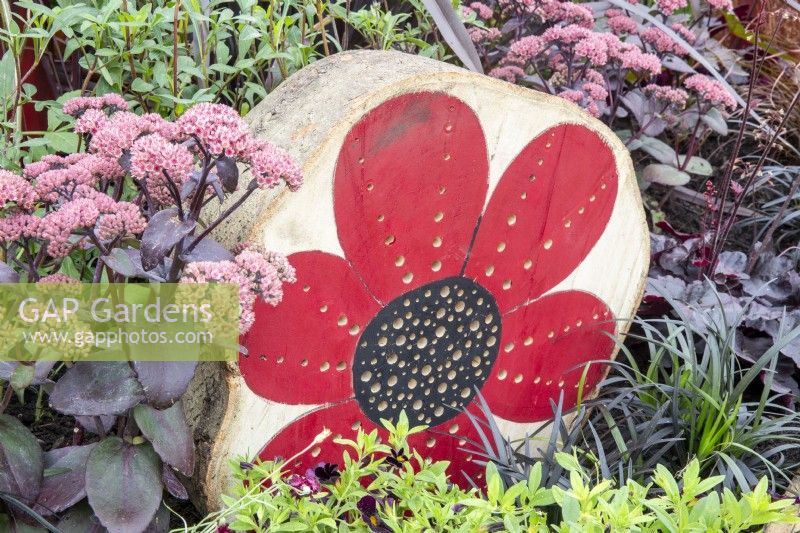 A tree stump made into an insect hotel with a painted poppy flower for decoration, planting of Sedum