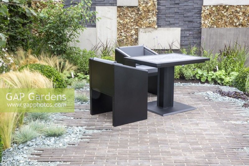 Brick paved patio area with black table and chairs, wall made from grey brick and concrete slabs - mixed perennial planting 