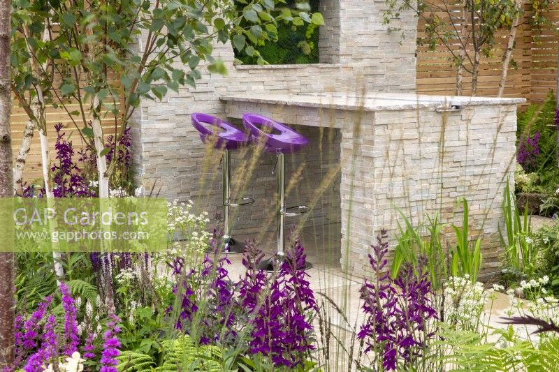 A stone dining area with purple bar stools, mixed perennial planting in purple and white colours