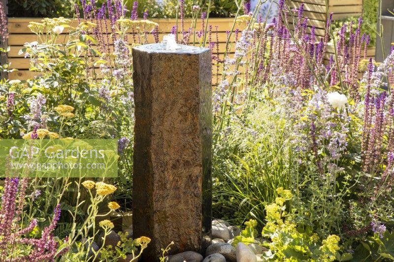 A stone column water feature with mixed perennial planting including Achillea millefolium and purple flowered Salvias