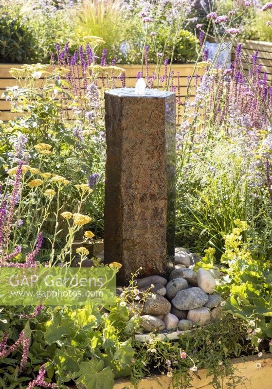 A stone column water feature with mixed perennial planting including Achillea millefolium and purple flowered Salvias