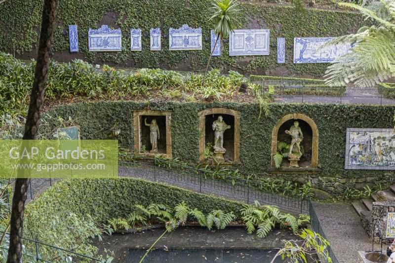 Several paths cross the steep sides of a valley with retaining walls, covered in ivy and with paths in front. On the bottom path, arched stone windows create niches with stone figurative statues in. A path above has antique tiled panels set on it. Monte Palace Gardens, Madeira. August. 