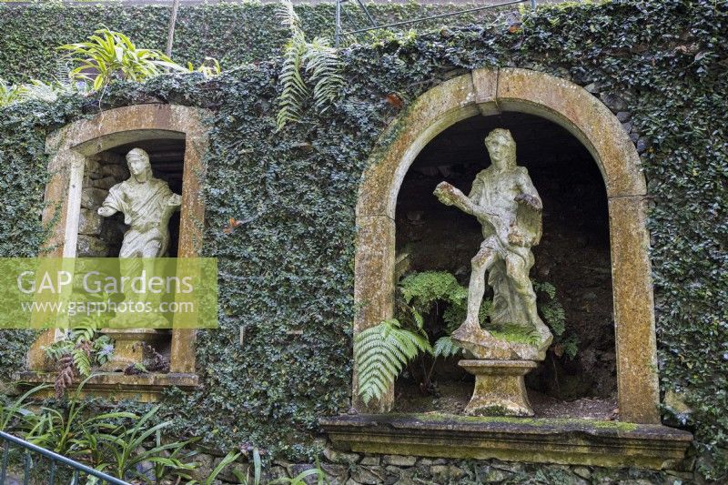 Old stone window arches set into ivy covered walls create niches for stone figurative statues. Monte Palace Gardens, madeira. August. 