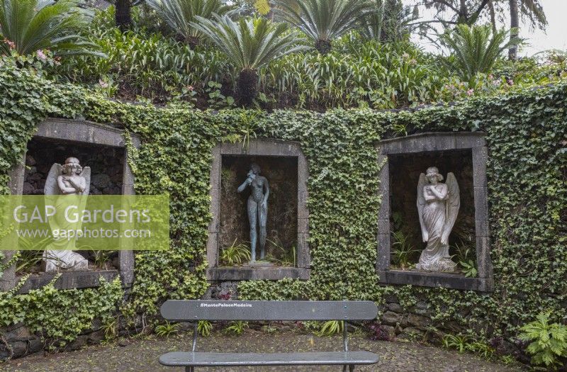 A curved ivy covered wall, with three stone window arches inset in the wall and figurative statues within the niches. A bench with the hashtag MontePalaceGardens is in the foreground. Monte Palace gardens, Madeira. August. 