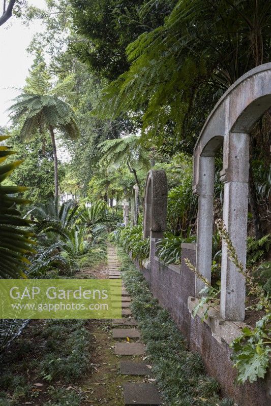 A stone slab path leads into the distance with arched stone windows beside it, set within lush tropical planting. Monte Palace Gardens, Madeira
