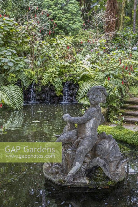A figurative statue in a pond, with small waterfalls in the background, steps to the right and lush tropical planting in the background. Monte Palace Gardens, Madeira
