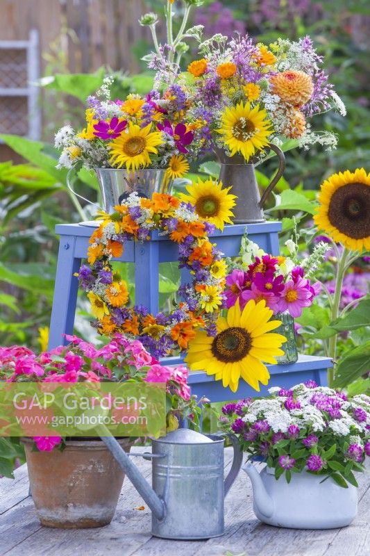 Floral display including Impatiens in terracotta pot, a wreath, teapot with red clover and yarrow and many bouquets in vases featuring Helianthus, Cosmos, Calendula, Tropaeolum, Dahlia and wildflowers.